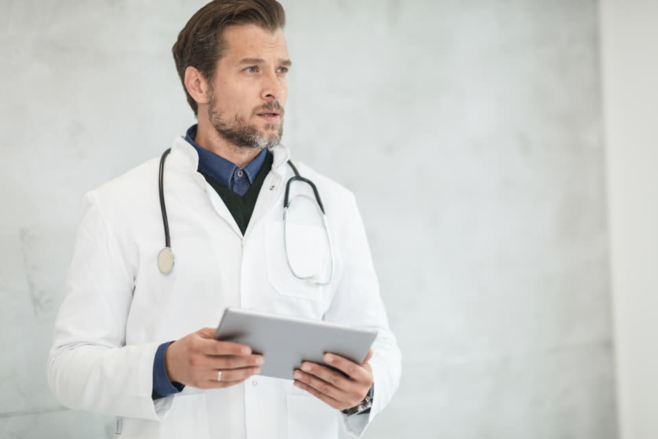 Dr. Daniel Reyes in a white coat with a tablet in front of a pale wall